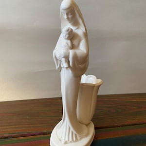 May include: A white ceramic figurine of a woman holding a baby. The woman is wearing a veil and a long gown. The figurine has a tulip-shaped vase attached to the side. The statue is approximately 8 inches tall.