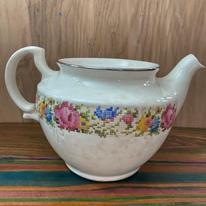 Op de afbeelding: Een witte keramische theepot met een zilveren rand en een gebogen handvat. De theepot heeft een kruissteek bloemenpatroon in roze, blauw, geel en groen. De tuit zit aan de rechterkant.