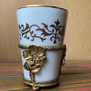 以下が含まれることがあります： 金色の花柄とトリムが施された白い磁器のカップ。カップは、前面に大きな花が付いた金色の金属ホルダーに収められています。カップの高さは約10cmです。