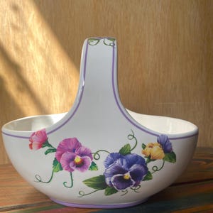 May include: A white ceramic basket with a high arched handle, decorated with painted pansy flowers in shades of purple, pink, and yellow. The rim and handle are accented with a thin purple line, and the base is trimmed with a matching purple band.