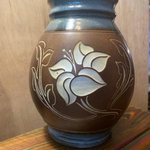 May include: A brown ceramic vase with a blue rim and base. The vase features a large, stylized flower design in cream and light blue, with matching leaf accents. The vase is a decorative piece.