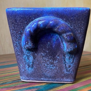 May include: A square, ceramic planter in shades of blue. The planter has a textured surface and a curved handle on the front. The handle is a lighter shade of blue than the rest of the planter. The planter is sitting on a colourful, striped surface.