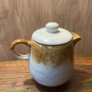 May include: A ceramic teapot with a rounded body and a lid. The teapot is primarily white with a brown gradient that darkens towards the top. The handle and spout are brown. The lid is white with small brown speckles.