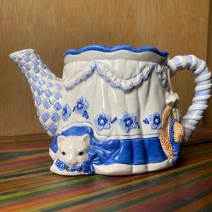 May include: A ceramic teapot with a white and blue design. The teapot features a cat peeking out from the front, blue floral accents, and a woven pattern on the spout. A small straw hat is attached to the side.