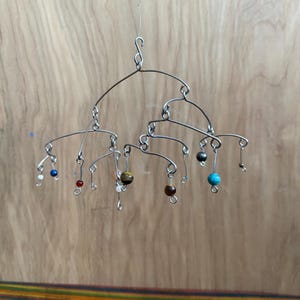 May include: A handcrafted mobile featuring a wire frame with various colorful beads. The beads are in different sizes and colors, including blue, red, brown, and white. The mobile hangs from a thin wire, creating a delicate and artistic display.