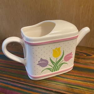 May include: Cream-colored ceramic watering can with a square body and a curved handle. The can features a pink trim, small gray polka dots, and a floral design of yellow and purple tulips. A decorative piece for gardening.