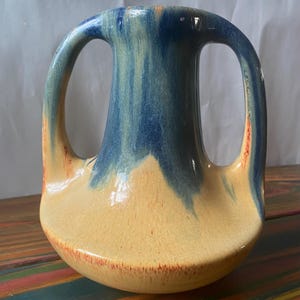 May include: A ceramic vase with two arched handles. The vase has a rounded base and a wide opening. The top is blue, transitioning to a tan colour, with orange accents. The vase is on a multi-coloured striped surface.