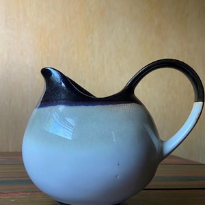 May include: A ceramic pitcher with a rounded body and a curved handle. The pitcher features a gradient of colors, transitioning from white to light blue, with a dark blue rim and handle. The handle has a white accent.