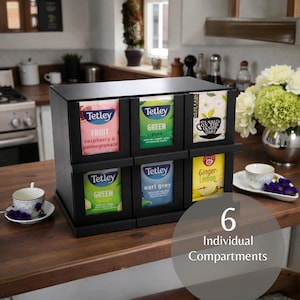 Può includere: Un organizer per bustine di tè in legno nero con sei scomparti individuali. Ogni scomparto contiene un diverso tè Tetley, tra cui frutta, verde e Earl Grey. Il testo "6 Scomparti Individuali" è visibile.
