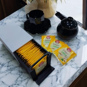 Könnte beinhalten: Ein Teeservice mit schwarzer Teekanne, Tasse und Untertasse. Eine weiße und schwarze Box enthält einzeln verpackte Teebeutel. Mehrere Ginger-Lemon Teebeutel liegen daneben, was auf ein Teetrinken hindeutet.