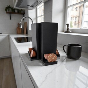 Könnte beinhalten: Ein schwarzer Kaffeepadhalter und eine schwarze Tasse auf einer weißen Arbeitsplatte. Der Halter hat Fächer zur Aufbewahrung von Kaffeepads. Die Küchenumgebung umfasst ein Waschbecken, einen Wasserhahn und ein Fenster.