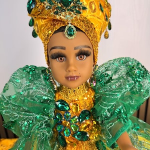 May include: A doll wearing a yellow headpiece adorned with green teardrop-shaped gems and gold sequins. The doll features brown eyes, long eyelashes, and a yellow dress with green sequined lace sleeves and a bodice decorated with large green gems.