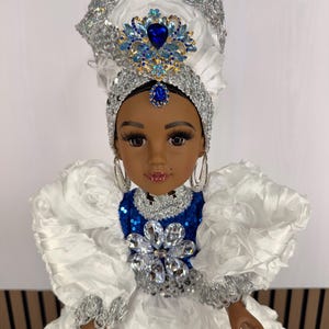 May include: A doll with dark skin and long eyelashes wearing a white ruffled dress with a blue sequined bodice. The doll features a large silver headpiece adorned with a blue and clear crystal brooch and a matching blue crystal forehead pendant.