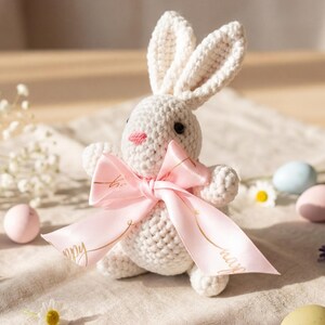 Puede incluir: Un conejito blanco tejido a ganchillo con un lazo rosa, ojos negros y una nariz rosa. El conejito está rodeado de huevos de Pascua de colores pastel y pequeñas flores blancas sobre una superficie de lino beige.