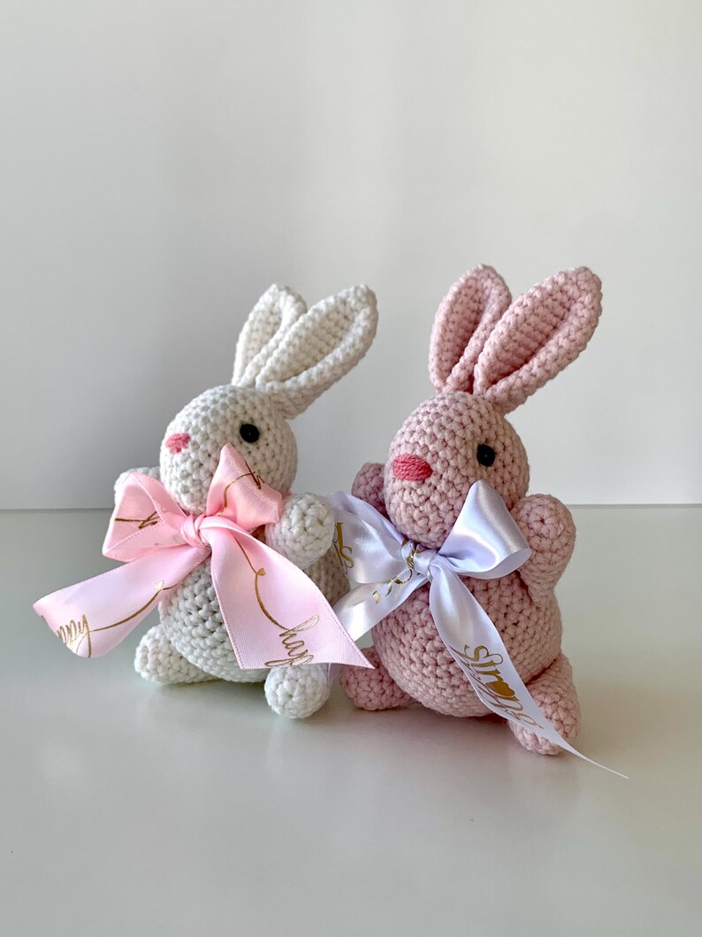 K&ouml;nnte beinhalten: Zwei geh&auml;kelte Hasen, einer wei&szlig; und einer rosa, werden gezeigt. Jeder Hase hat eine Schleife aus Satinband um den Hals. Der wei&szlig;e Hase hat eine rosa Schleife, und der rosa Hase hat eine wei&szlig;e Schleife. Die Hasen haben schwarze Augen und rosa Nasen.