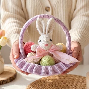 May include: A light purple crocheted Easter basket, filled with colourful crocheted eggs and a white bunny with a pink bow. The basket has a handle and a ruffled edge. A pink ribbon with gold text is wrapped around the basket.
