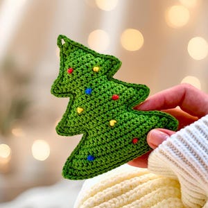 May include: A green crocheted Christmas tree ornament with colorful bead accents. The handmade tree is held against a soft, blurred background. The tree is approximately 4 inches tall.