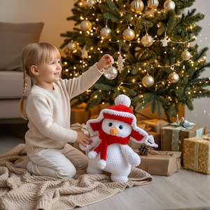 Puede incluir: Una niña decora un árbol de Navidad con adornos. Un muñeco de nieve blanco hecho a mano con gorro y bufanda rojos está cerca. El árbol está adornado con adornos y luces doradas, y hay regalos envueltos en la base.