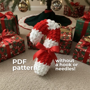 Puede incluir: Un bastón de caramelo de ganchillo rojo y blanco hecho a mano con un lazo de satén rojo. El bastón de caramelo se coloca frente a regalos de Navidad envueltos y un árbol de Navidad. El texto "PDF pattern" y "without a hook or needles!" son visibles.