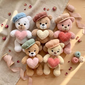 Pode incluir: Uma coleção de ursinhos de pelúcia de crochê feitos à mão, cada um com um detalhe em forma de coração. Os ursinhos são em várias cores, incluindo branco, bege e rosa, e usam boinas. O fundo inclui pétalas de rosa secas e carretéis de linha.