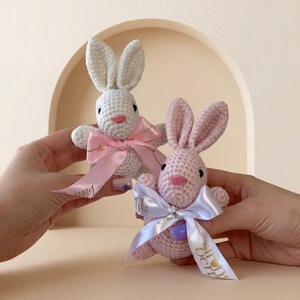 K&ouml;nnte beinhalten: Zwei geh&auml;kelte Hasen, einer wei&szlig; und einer rosa, jeweils mit einer rosa Schleife und einem Band. Die Hasen haben schwarze Augen und rosa Nasen. Der wei&szlig;e Hase hat ein rosa Band, und der rosa Hase hat ein wei&szlig;es Band.