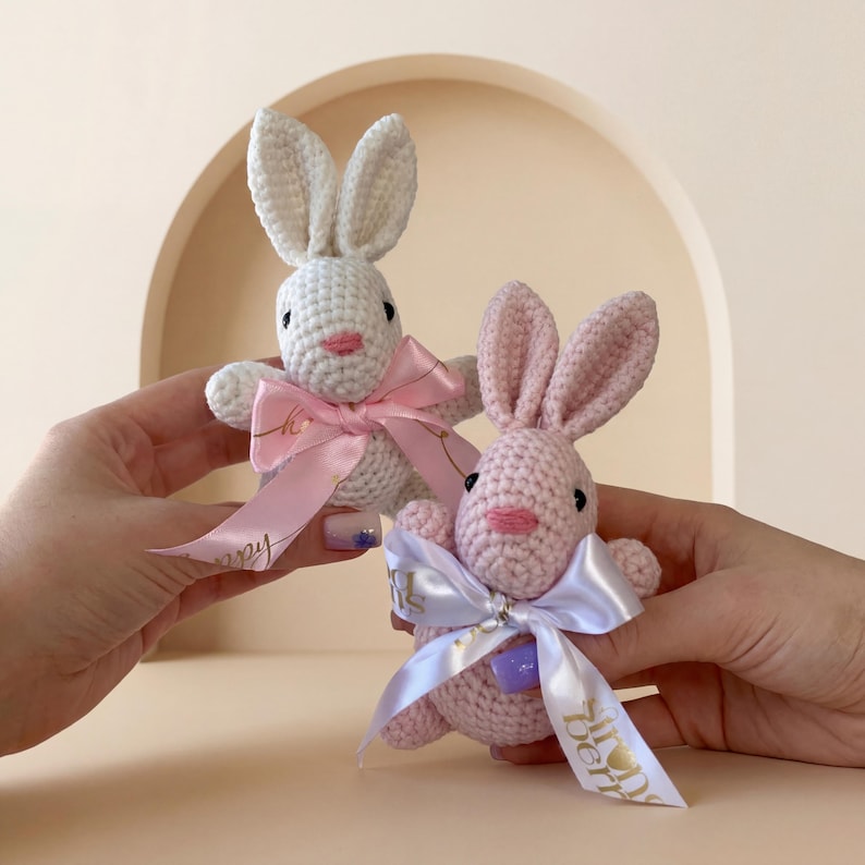 K&ouml;nnte beinhalten: Zwei geh&auml;kelte Hasen, einer wei&szlig; und einer rosa, jeweils mit einer rosa Schleife und einem Band. Die Hasen haben schwarze Augen und rosa Nasen. Der wei&szlig;e Hase hat ein rosa Band, und der rosa Hase hat ein wei&szlig;es Band.
