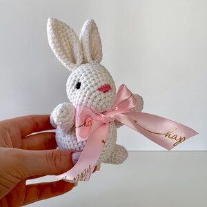 K&ouml;nnte beinhalten: Ein handgefertigtes wei&szlig;es H&auml;schen aus H&auml;kelarbeit mit einer rosa Schleife. Das H&auml;schen hat schwarze Augen und einen rosa Mund. Das rosa Band hat einen goldenen Text mit der Aufschrift "happy". Das H&auml;schen wird in der Hand einer Person gehalten.