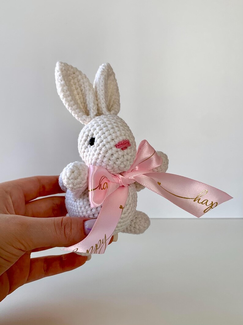 K&ouml;nnte beinhalten: Ein handgefertigtes wei&szlig;es H&auml;schen aus H&auml;kelarbeit mit einer rosa Schleife. Das H&auml;schen hat schwarze Augen und einen rosa Mund. Das rosa Band hat einen goldenen Text mit der Aufschrift "happy". Das H&auml;schen wird in der Hand einer Person gehalten.