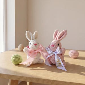 K&ouml;nnte beinhalten: Zwei geh&auml;kelte Osterhasen, einer wei&szlig; und einer rosa, sind mit rosa und lavendelfarbenen B&auml;ndern verziert. Ein gr&uuml;ner geh&auml;kelter Ball und mehrere geh&auml;kelte Eier befinden sich auf einer Holzoberfl&auml;che. Die Hasen haben schwarze Augen und rosa Nasen.