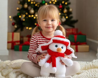 May include: A white snowman plush toy with a red hat and scarf. The snowman has blue eyes and an orange nose. The plush toy is being held by a child wearing a red and white striped top. A Christmas tree is in the background.