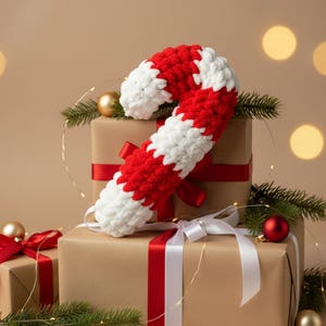 May include: A large, handmade, red and white striped crochet candy cane rests on wrapped gifts with red and white ribbons. The gifts are wrapped in brown paper. Christmas ornaments and greenery complete the festive scene.