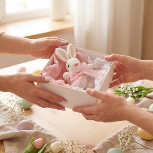 K&ouml;nnte beinhalten: Eine wei&szlig;e Geschenkbox, gehalten von zwei H&auml;nden, enth&auml;lt einen geh&auml;kelten Hasen mit rosa Schleife. Die Box ist mit rosa und wei&szlig;em Seidenpapier ausgekleidet. Ostereier und Blumen sind auf dem Holztisch verstreut.