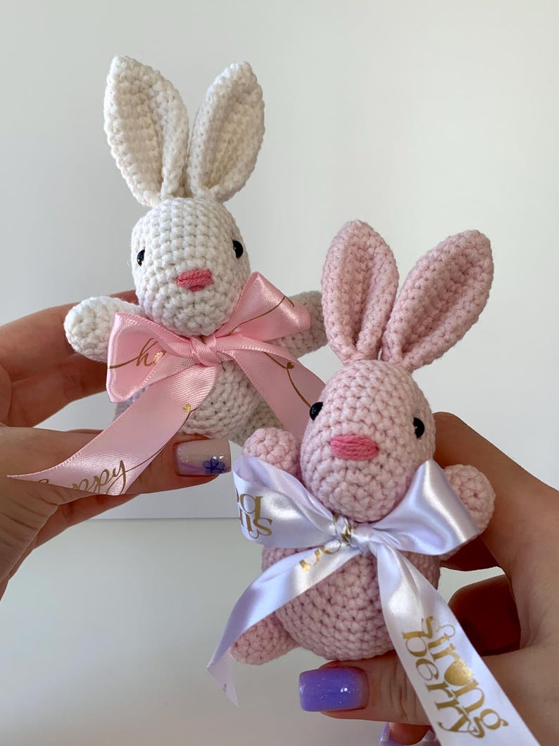 K&ouml;nnte beinhalten: Zwei geh&auml;kelte Hasen, einer wei&szlig; und einer rosa, werden vor einem wei&szlig;en Hintergrund pr&auml;sentiert. Jeder Hase hat lange Ohren, eine rosa Nase und eine Schleife aus Satinband. Der wei&szlig;e Hase hat eine rosa Schleife, und der rosa Hase hat eine wei&szlig;e Schleife.