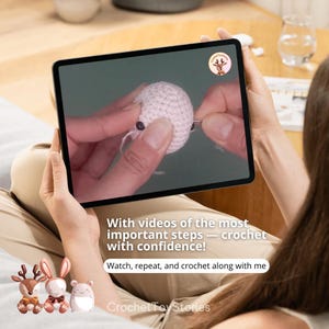 K&ouml;nnte beinhalten: Eine Person h&auml;lt ein Tablet mit einem H&auml;keltutorial. Der Bildschirm zeigt H&auml;nde, die an einem kleinen, wei&szlig;en geh&auml;kelten Objekt arbeiten. Der Text auf dem Bildschirm lautet: "Mit Videos der wichtigsten Schritte - h&auml;keln mit Zuversicht!"