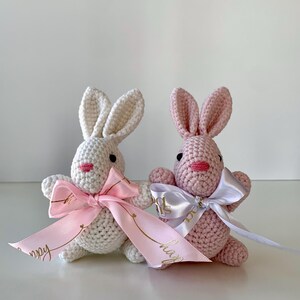 K&ouml;nnte beinhalten: Zwei geh&auml;kelte Hasen, einer wei&szlig; und einer rosa, mit rosa Schleifen und B&auml;ndern. Die B&auml;nder haben goldenen Text. Die Hasen haben schwarze Augen und rosa Nasen und M&uuml;nder. Die Hasen sitzen auf einer wei&szlig;en Oberfl&auml;che.
