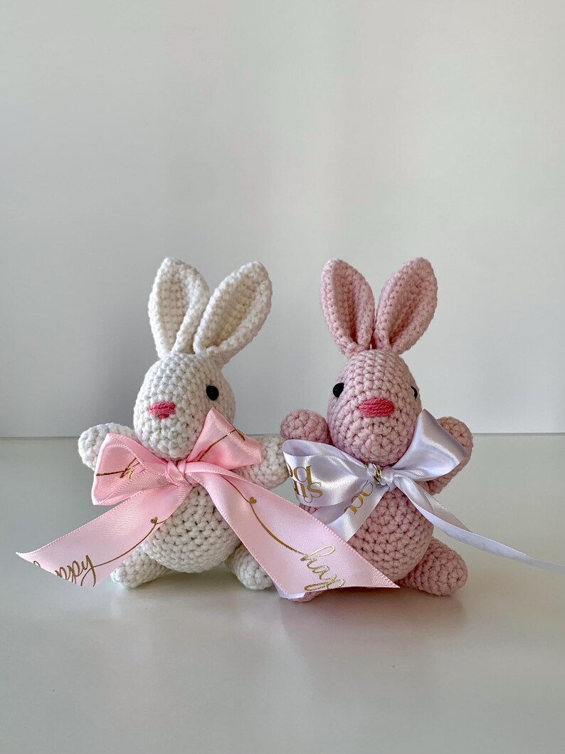 K&ouml;nnte beinhalten: Zwei geh&auml;kelte Hasen, einer wei&szlig; und einer rosa, mit rosa Schleifen und B&auml;ndern. Die B&auml;nder haben goldenen Text. Die Hasen haben schwarze Augen und rosa Nasen und M&uuml;nder. Die Hasen sitzen auf einer wei&szlig;en Oberfl&auml;che.