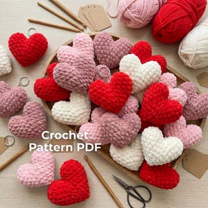May include: A collection of crocheted heart-shaped keychains in various shades of red, pink, and white, arranged on a wooden tray. The text "Crochet Pattern PDF" is visible. Knitting needles, yarn, and tags are also present.