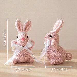 K&ouml;nnte beinhalten: Zwei rosa geh&auml;kelte Hasen mit wei&szlig;en Schleifen. Die Hasen sind 11 cm hoch und 7 cm und 7,5 cm lang. Die Hasen haben schwarze Augen und rosa Nasen. Die Hasen sitzen auf einer Holzoberfl&auml;che.