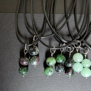 Pode incluir: Uma coleção de colares com cordões pretos e pingentes de pedras preciosas. Os pingentes apresentam várias cores, incluindo verde, rosa e preto, com um acabamento polido. Os colares são exibidos em um fundo escuro e texturizado.