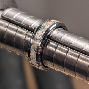 May include: A dark metallic ring featuring a central inlay composed of crushed white and tan stone fragments interspersed with small, iridescent green flecks. The ring is positioned on a metal ring mandrel used for sizing.