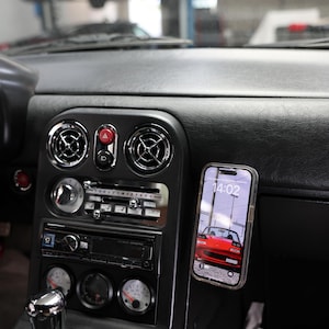 Puede incluir: Interior de coche con salpicadero negro y detalles cromados. Un smartphone muestra una imagen de coche rojo. El salpicadero incluye rejillas de ventilación, indicadores y un equipo de música. Un coche blanco es visible en el fondo.