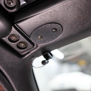 Puede incluir: Primer plano del interior negro de un coche, centrado en el revestimiento del techo. Se ven tornillos, un pequeño panel ovalado y un micrófono. La superficie texturizada y los detalles de hardware sugieren un diseño funcional.