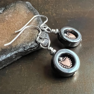 May include: A pair of dangle earrings featuring a dark gray circular design with a copper-colored heart-shaped center. The earrings have silver-colored ear wires and are displayed on a dark, textured surface.