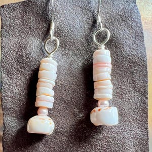 May include: A pair of dangle earrings featuring silver-toned metal hooks. Each earring consists of a vertical stack of pale pink, disc-shaped shell beads, a single small round pink bead, and a larger, irregular, smooth white shell pendant at the base.