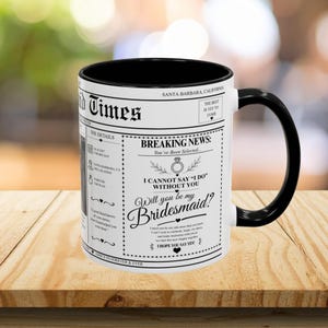 Puede incluir: Una taza de cerámica blanca con interior y asa negros. La taza presenta un diseño de estilo periódico con el titular "Breaking News: You've Been Selected..." y la pregunta "Will you be my Bridesmaid?"