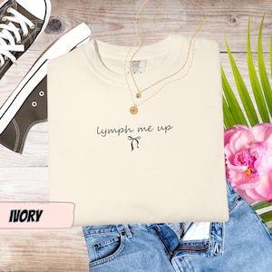 Könnte beinhalten: Elfenbeinfarbenes T-Shirt mit dem Text "lymph me up" und einer Schleifengrafik. Das Shirt wird mit einer goldenen Halskette, einer blauen Jeans und einem Paar schwarzer Turnschuhe kombiniert. Eine rosa Blume und grüne Blätter sind ebenfalls im Bild.