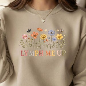 Könnte beinhalten: Beiges Sweatshirt mit farbenfrohem Blumenmuster und dem Text "LYMPH ME UP". Die Blumen sind in Rosa, Orange, Gelb und Blau gehalten, mit grünen Blättern und Schmetterlingen.
