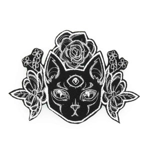 May include: Black and white embroidered patch of a cat with a third eye and flowers. The cat's eyes are outlined in white and the flowers are detailed with white stitching.