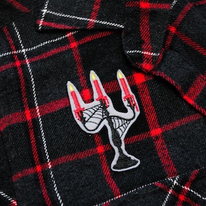 May include: A black and red plaid flannel shirt with a black and white embroidered patch of a candelabra with three lit candles. The patch is on the left chest pocket of the shirt.