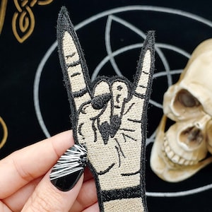 Może przedstawiać: Beige naszywka do prasowania z haftem przedstawiającym rękę wykonującą gest rock and roll. Naszywka znajduje się na czarnym tle z złotym wzorem celtyckiego węzła.